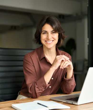 Profissional sorridente sentado à secretária, com portátil e caderno, em ambiente de trabalho